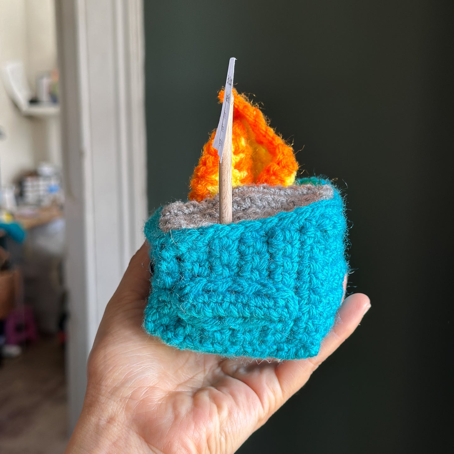 Hand holding a small crocheted dunpster with a flame on a dark background