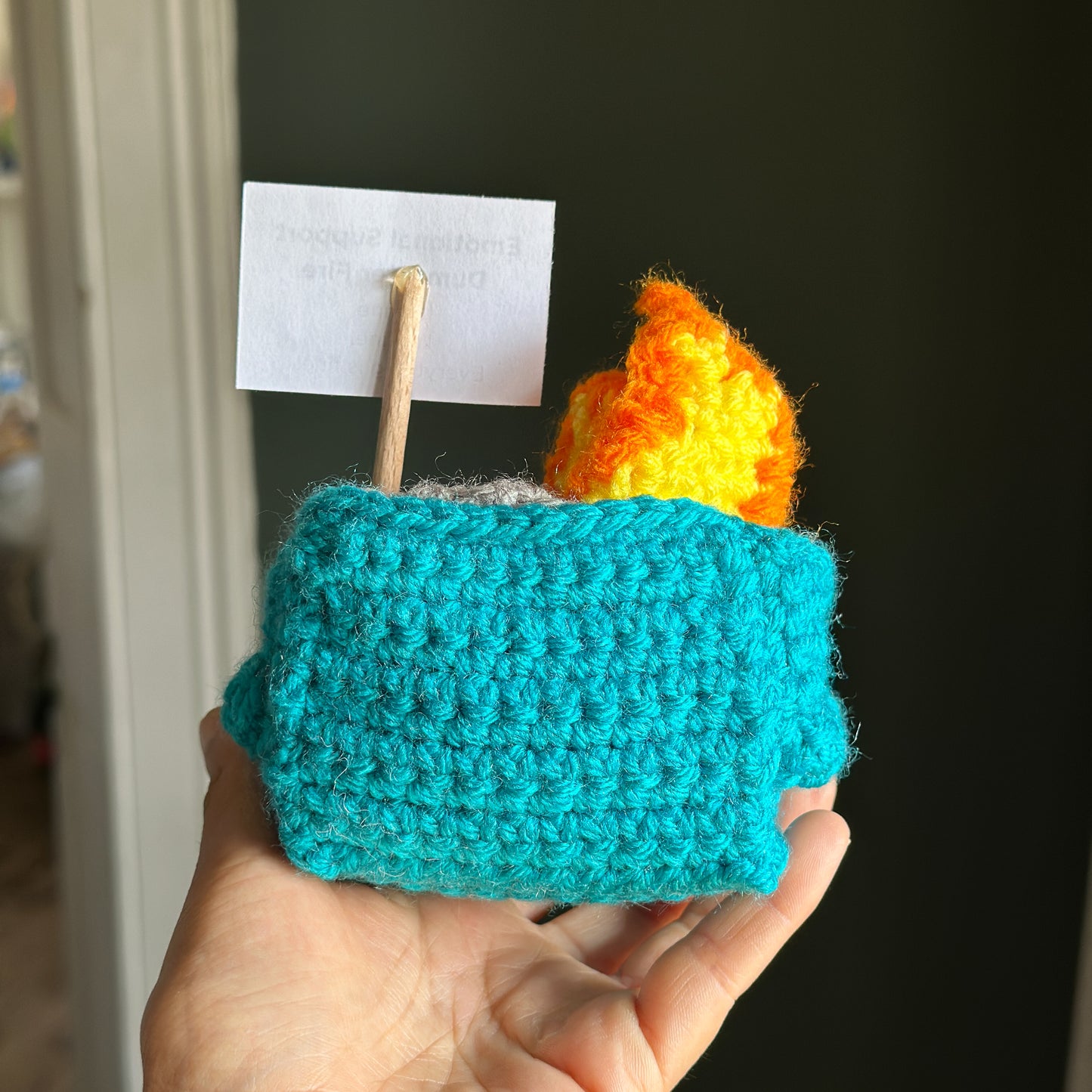 Hand holding a teal crocheted dumpster item with orange and yellow flames against a dark background