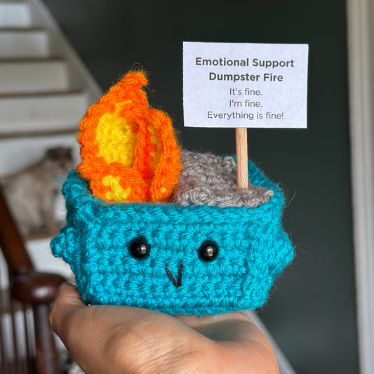 Crocheted emotional support dumpster fire with a flame and sign held by a stick on the top.