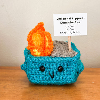 Crocheted emotional support dumpster fire with flames and text on a wooden surface.