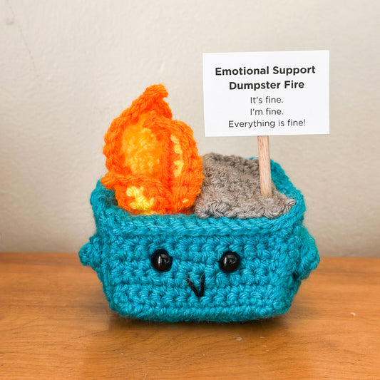Crocheted emotional support dumpster fire with flames and text on a wooden surface.