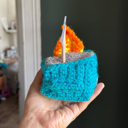 Hand holding a small crocheted dunpster with a flame on a dark background