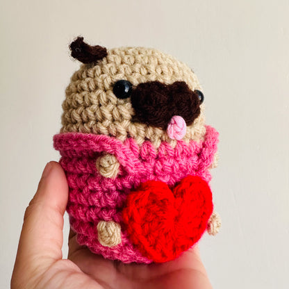 Handmade crochet pug toy wearing a pink overall with a red heart on a plain background