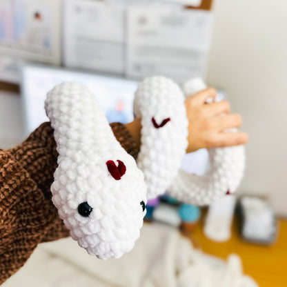 White snake plush toy with a red heart and  black safety eyes on a blurred indoor background