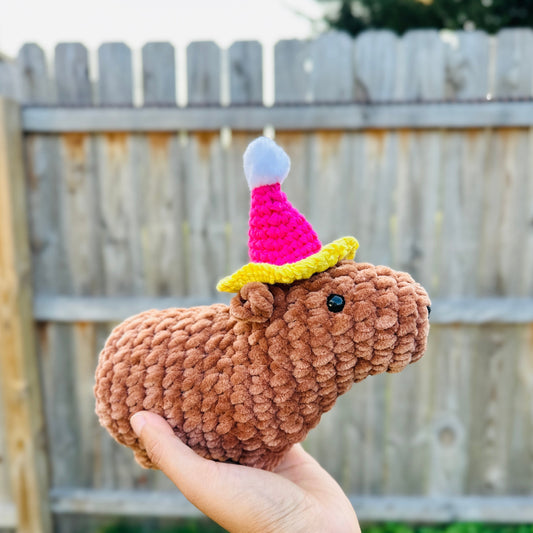 crochet capybara wearing a pink birthday hat in front of a fenced yard